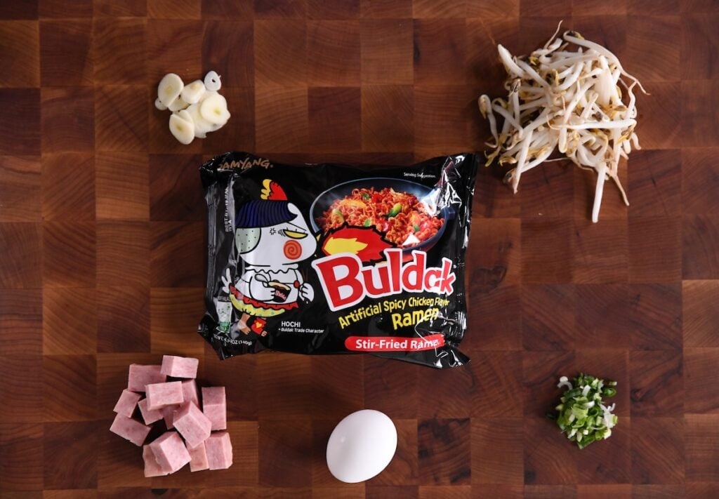 sliced garlic, beansprouts, green onions, egg, diced spam, and a package of buldak noodles on a cutting board