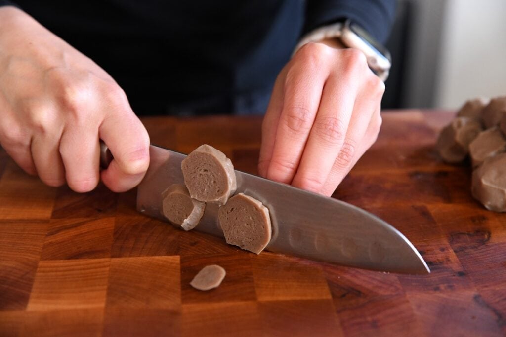 A close-up view of a home cook’s hands using a sharp chef's knife to slice a firm Vietnamese beef meatball into thin discs on a dark wood cutting board.