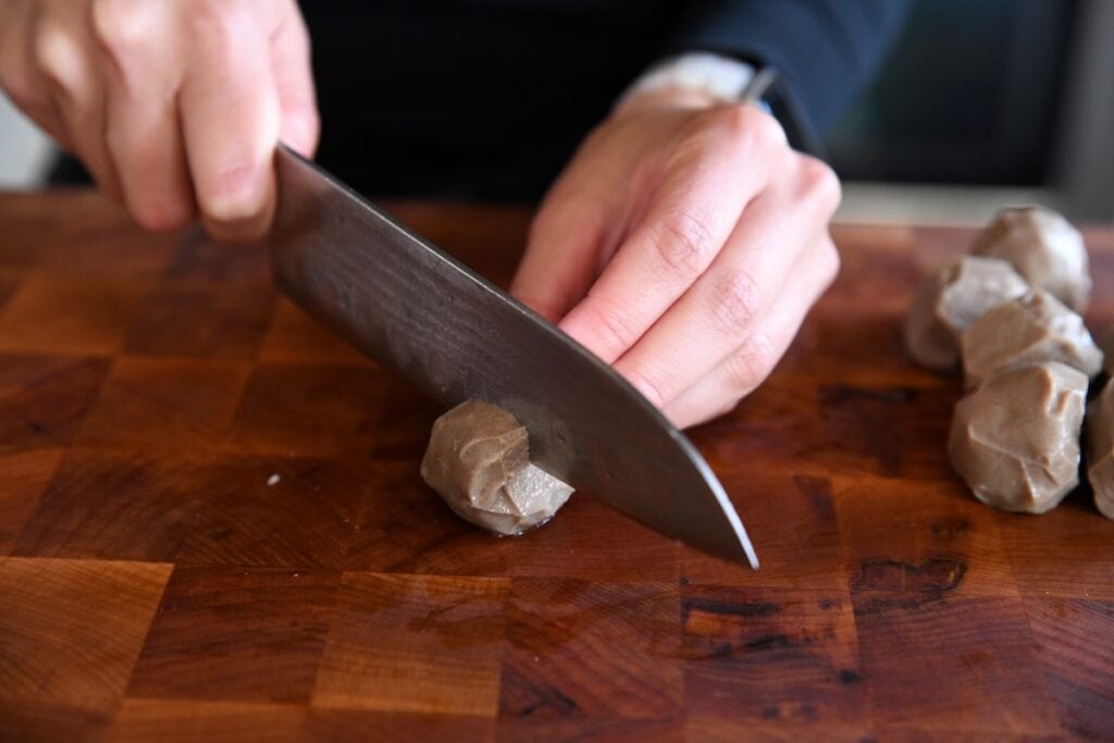 The hand of a cook carefully slicing a dense Vietnamese bò viên (beef meatball) in half on a rich, dark wood cutting board.
