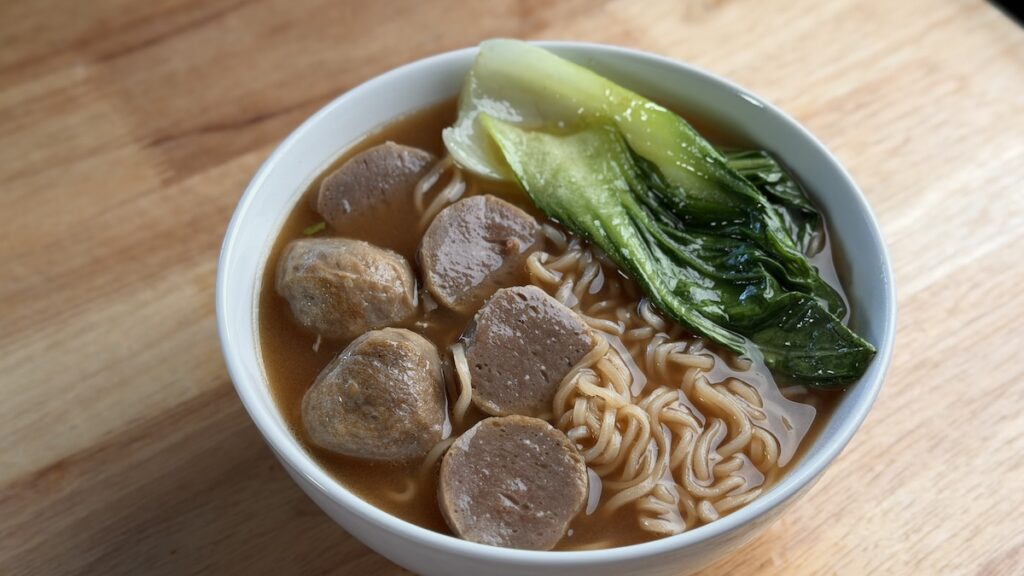 A bowl of Sapporo Ichiban beef ramen topped with sliced Vietnamese beef meatballs, a whole leaf of green bok choy, and fresh scallions in a savory brown broth.