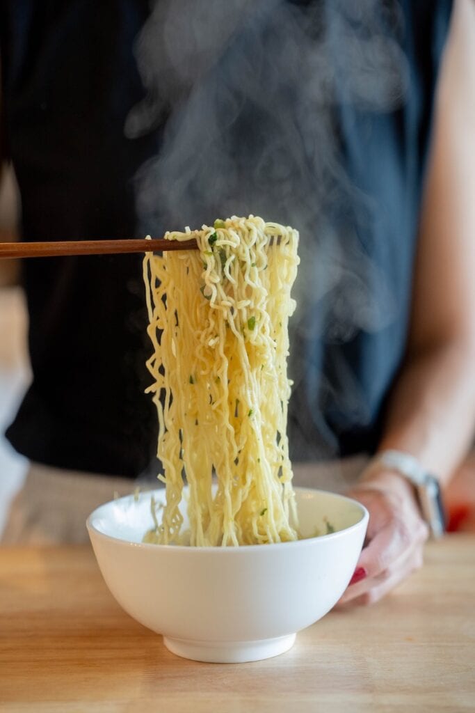 A close-up of the Kang Shi Fu Beef Instant Noodles with Sichuan Green Pepper being lifted from a white bowl with wooden chopsticks, showing wavy pale-yellow wheat noodles glossy with broth, small green scallion specks, and steam rising above them.