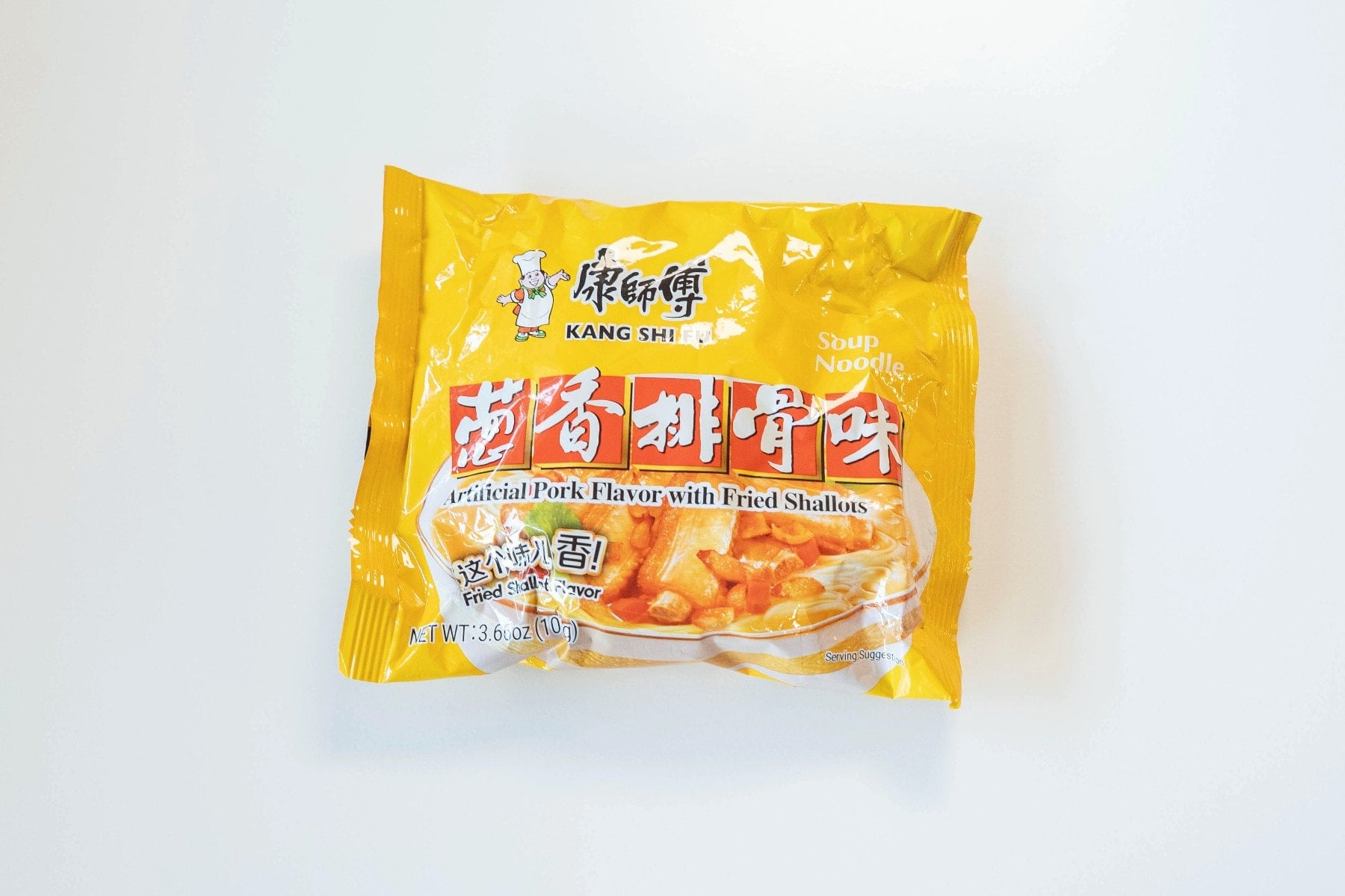 The bright yellow package of Kang Shi Fu Pork Instant Noodles with Shallots on a white surface, showing the small white-hatted Kang Shi Fu chef logo at the top, ๅบทๅธๅ
/ KANG SHI FU in red, ่ฑ้ฆๆ้ชจๅณ (Scallion Pork Rib Flavor) in white kanji on red blocks, "Artificial Pork Flavor with Fried Shallots" in white English text, "Soup Noodle" in the upper right, and a photo of finished noodles with sliced pork ribs and red bell pepper bits in a cream-colored broth.