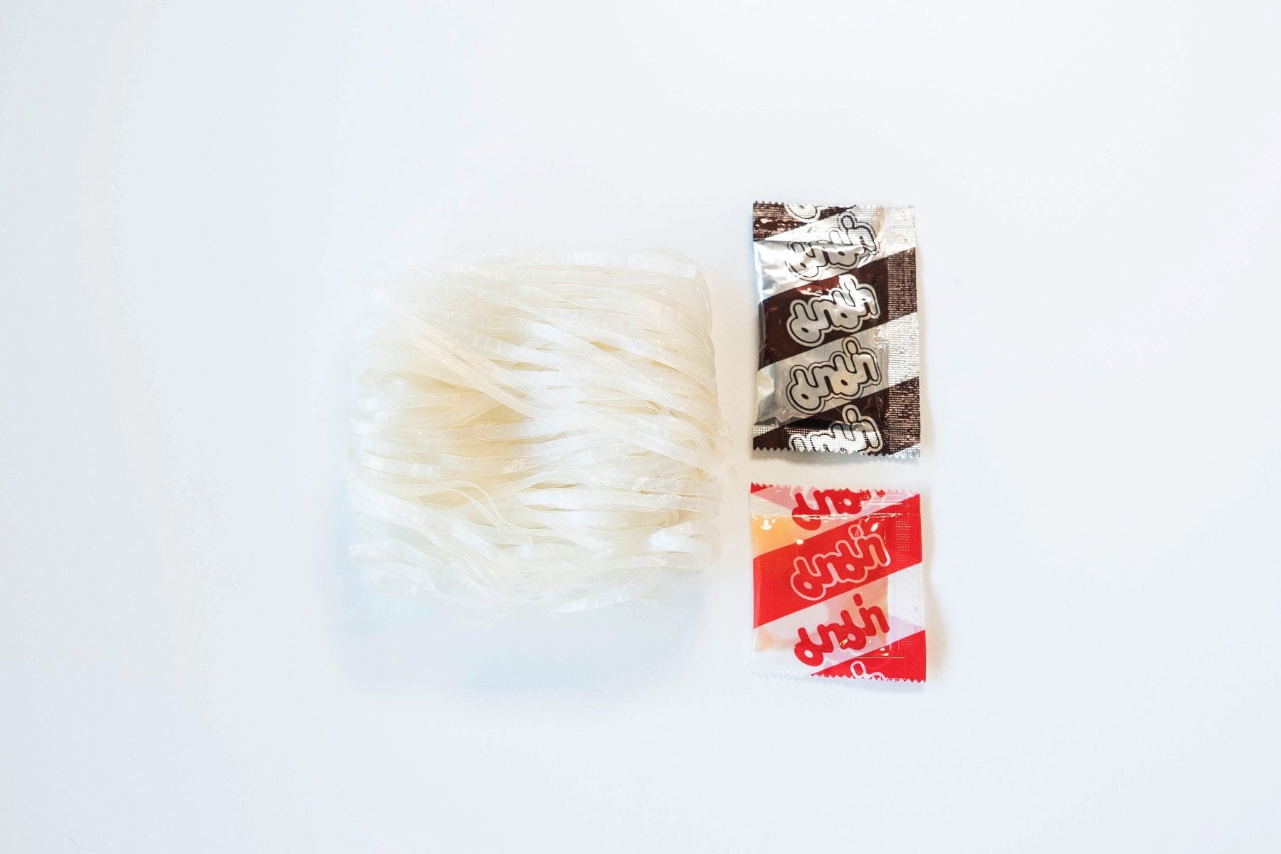 The contents of the MAMA Phแป Bรฒ Beef Noodle Soup pack laid out on a white surface, showing a folded bundle of thin flat white rice noodles on the left, a small black-and-silver MAMA seasoning powder sachet on the top right, and a small red sauce/oil packet on the bottom right.