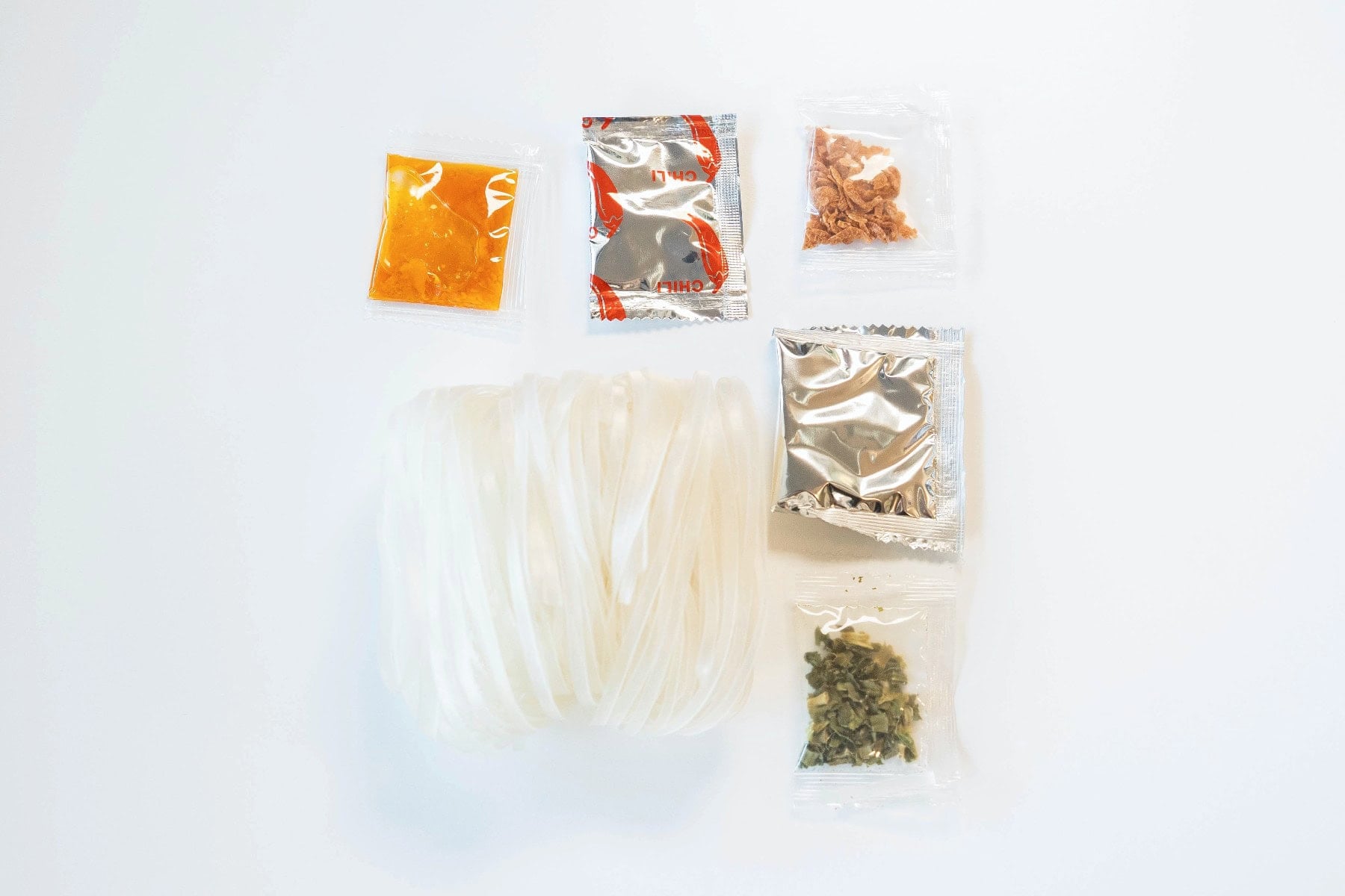 The contents of the Nguyen's Pho Instant Rice Noodles Seafood Flavor cup laid out on a white surface. A bundle of flat rice vermicelli noodles sits next to five small packets: an orange chili sauce, a silver-and-red chili powder, a clear packet of dried shrimp bits, a silver foil seasoning packet, and a small packet of dried green herbs
