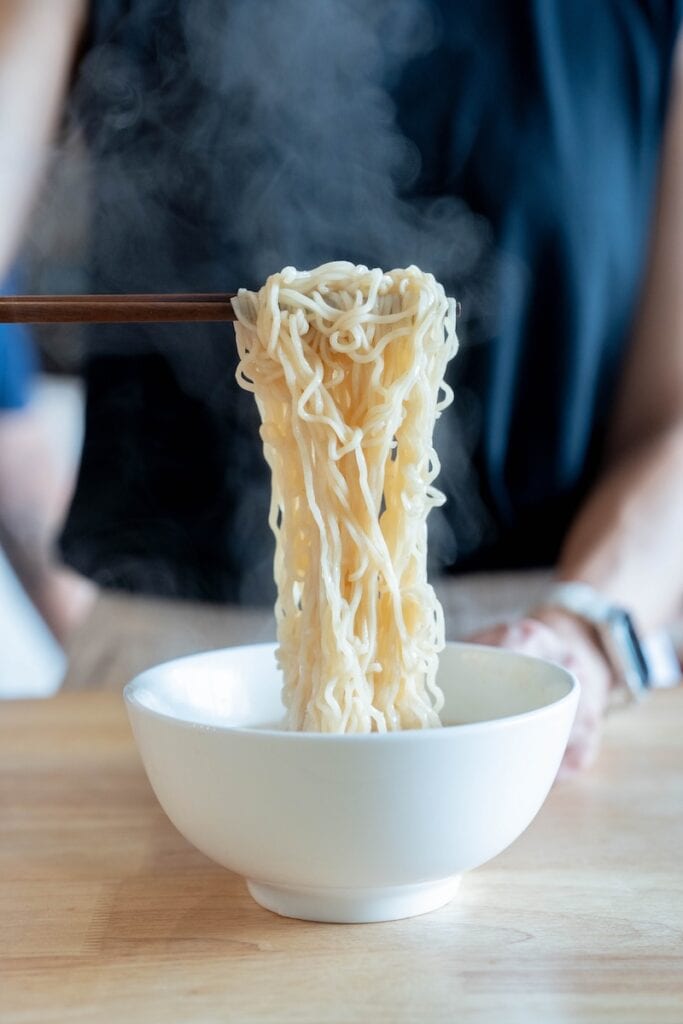 A close-up of the Sapporo Ichiban Hot & Spicy Chicken noodles being lifted from a white bowl with wooden chopsticks, steam rising, showing the brand's familiar wavy wheat noodles dripping with pale broth.