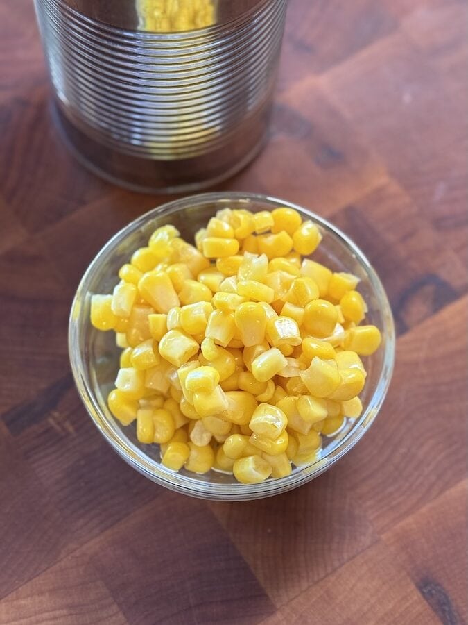 A small glass bowl filled with sweet yellow canned corn kernels on a dark wood grain cutting board.