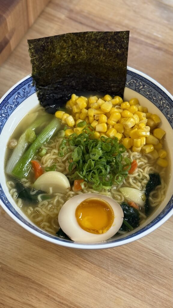 A bowl of instant ramen featuring a soft-boiled egg, nori seaweed, green onions, and a large portion of sweet canned corn.