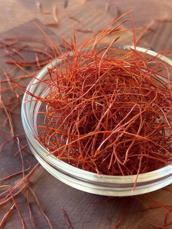 A small glass ramekin overflowing with bright red dried chili threads on a dark wooden cutting board.
