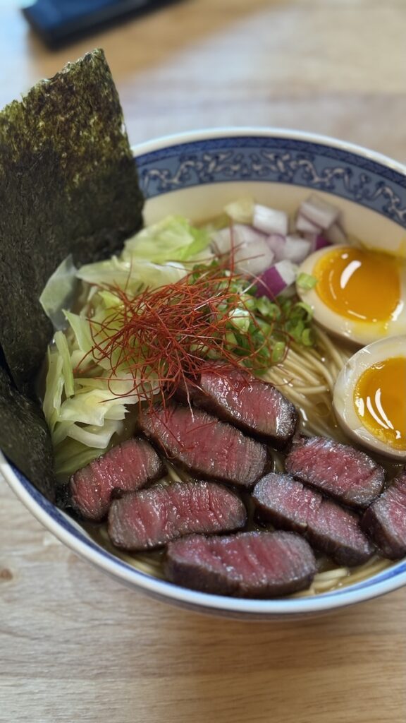 Instant ramen topped with perfectly seared medium-rare ribeye steak slices, jammy eggs, nori, red onions, and a garnish of delicate red chili threads