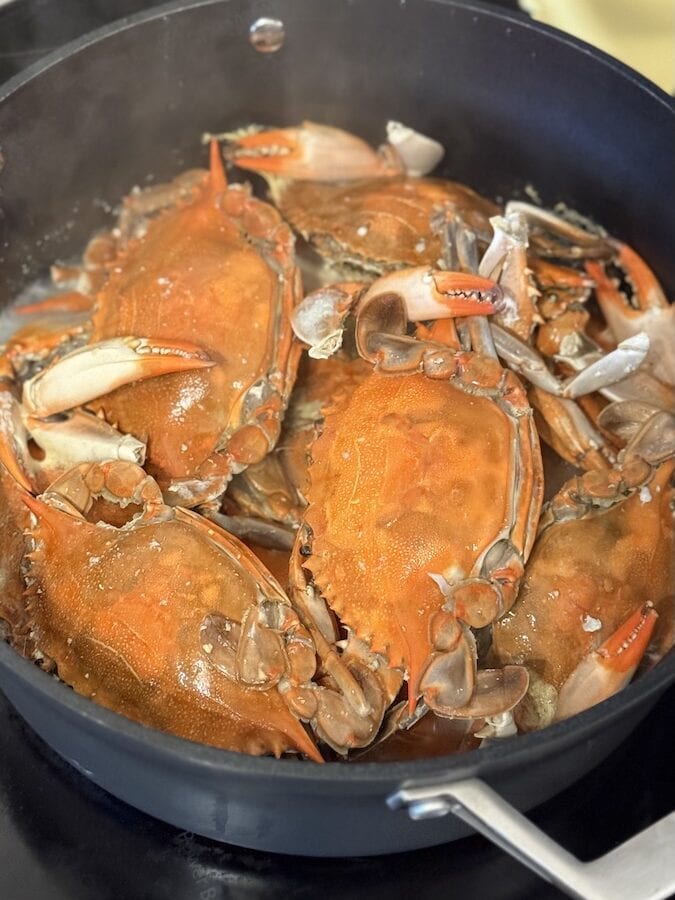 Several whole, bright orange steamed blue crabs being heated in a large black non-stick pot on a glass stovetop.