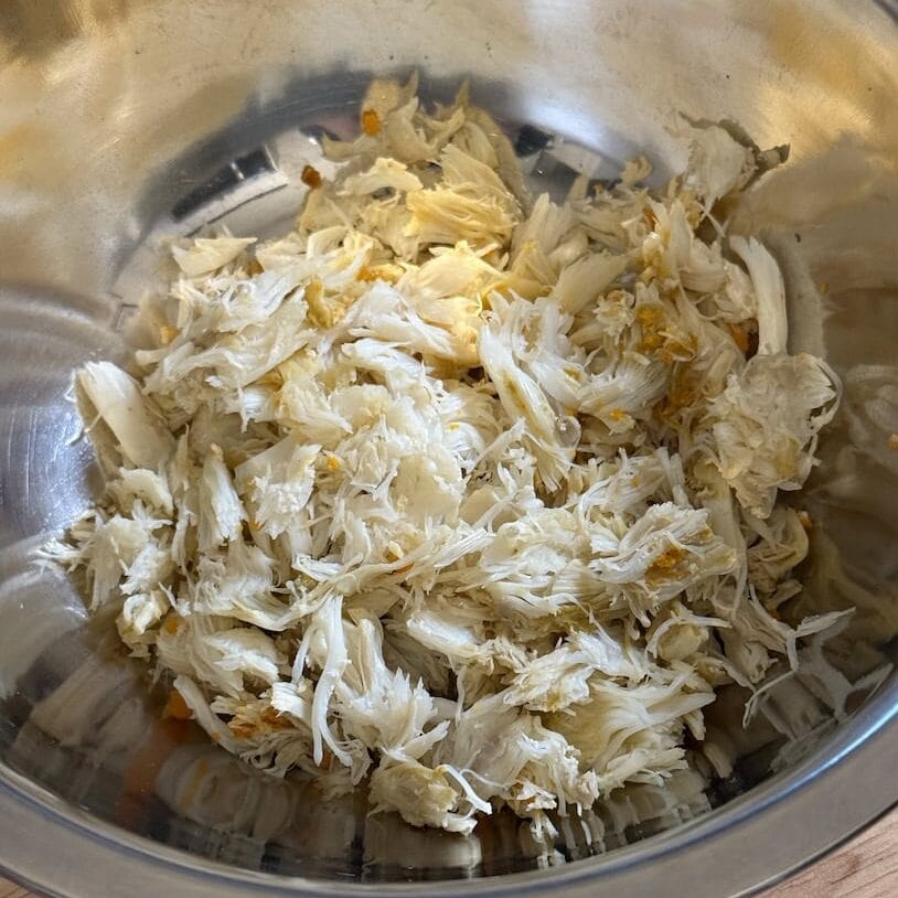 A stainless steel mixing bowl filled with freshly picked, shredded white crab meat sitting on a wooden countertop.