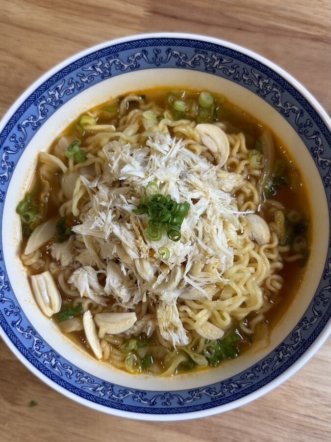 A blue and white patterned bowl of spicy instant ramen topped with a generous mound of fresh shredded crab meat, sliced raw garlic, and chopped green onions.