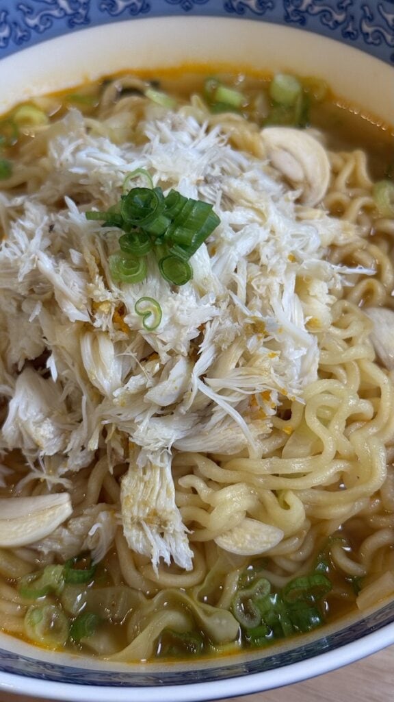 A macro close-up of a ramen bowl highlighting the texture of thick, wavy noodles and a mountain of fresh white crab meat garnished with vibrant green onions.