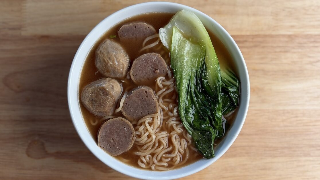 An overhead view of a bowl of instant ramen in a rich beef broth, topped with several hearty beef meatballs and a whole, vibrant green piece of blanched bok choy, served on a light wood grain table.