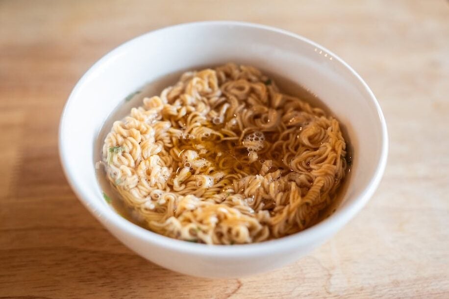 A white bowl of Hao Hao noodles steeping in hot water with visible oil from the seasoning packet on the surface.