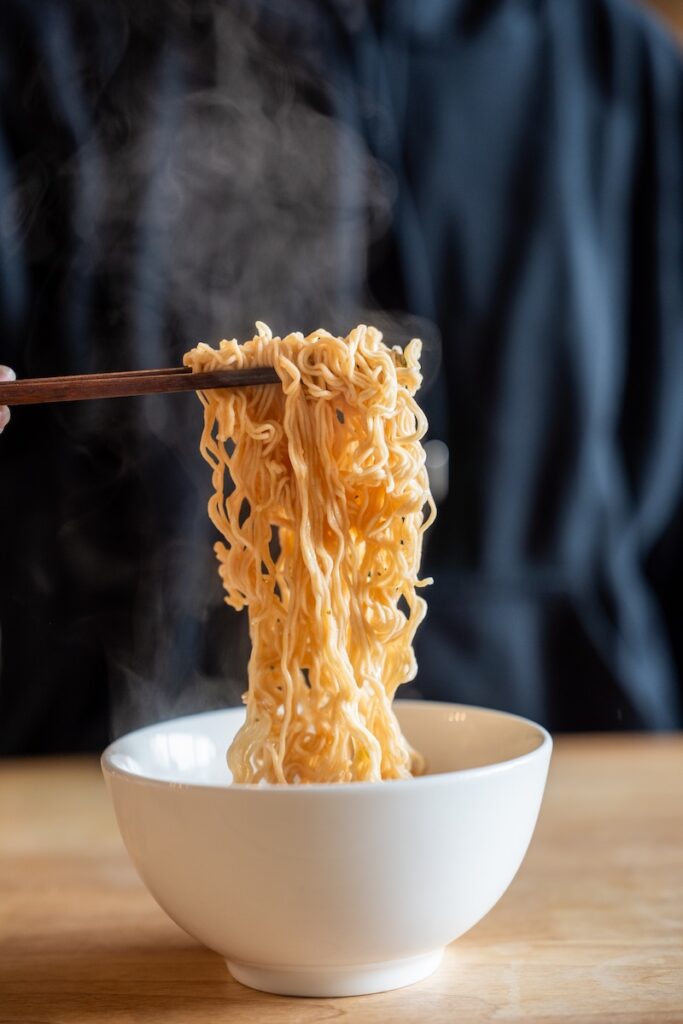 A close-up shot of steaming Hao Hao noodles being lifted from a white bowl with wooden chopsticks.
