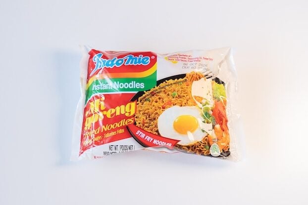 The red, white, and rainbow-striped package of Indomie Mi Goreng Instant Stir Fry Noodles on a white surface, showing the Indomie logo at top left, a photo of finished fried noodles with a sunny-side-up egg and vegetables on the right, and Mi Goreng FRIED NOODLES in red script.