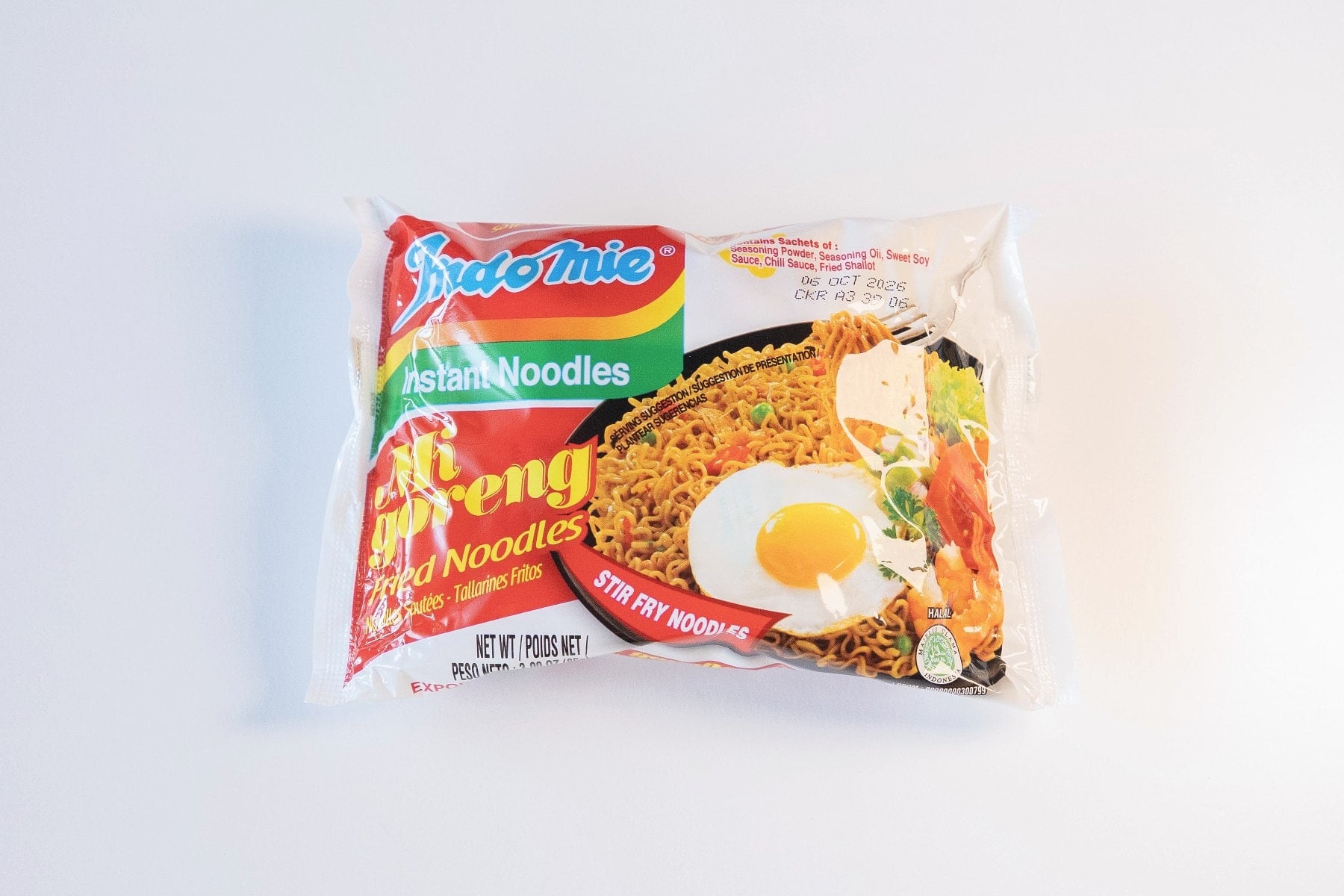 The red, white, and rainbow-striped package of Indomie Mi Goreng Instant Stir Fry Noodles on a white surface, showing the Indomie logo at top left, a photo of finished fried noodles with a sunny-side-up egg and vegetables on the right, and Mi Goreng FRIED NOODLES in red script.