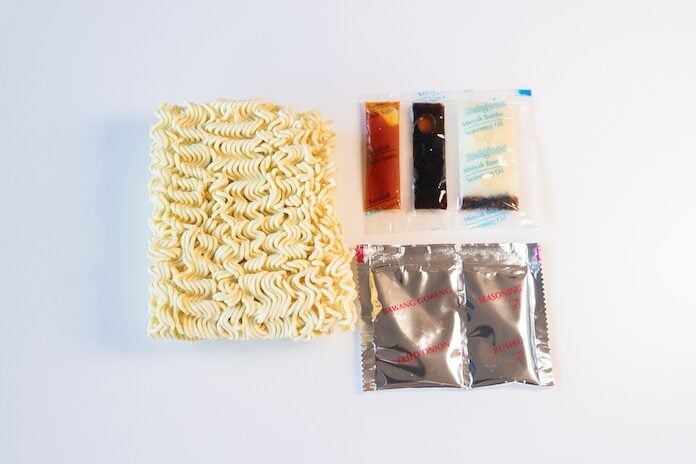 The contents of the Indomie Mi Goreng Instant Stir Fry Noodles pack laid out on a white surface, showing a rectangular block of thin wavy wheat noodles on the left with five sachets to the right. Three small horizontal sachets at top (orange, dark brown, clear) for the seasoning oil, sweet soy sauce, and chili sauce, and two silver foil sachets at the bottom labeled BAWANG GORENG (fried shallots) and SEASONING (bumbu).