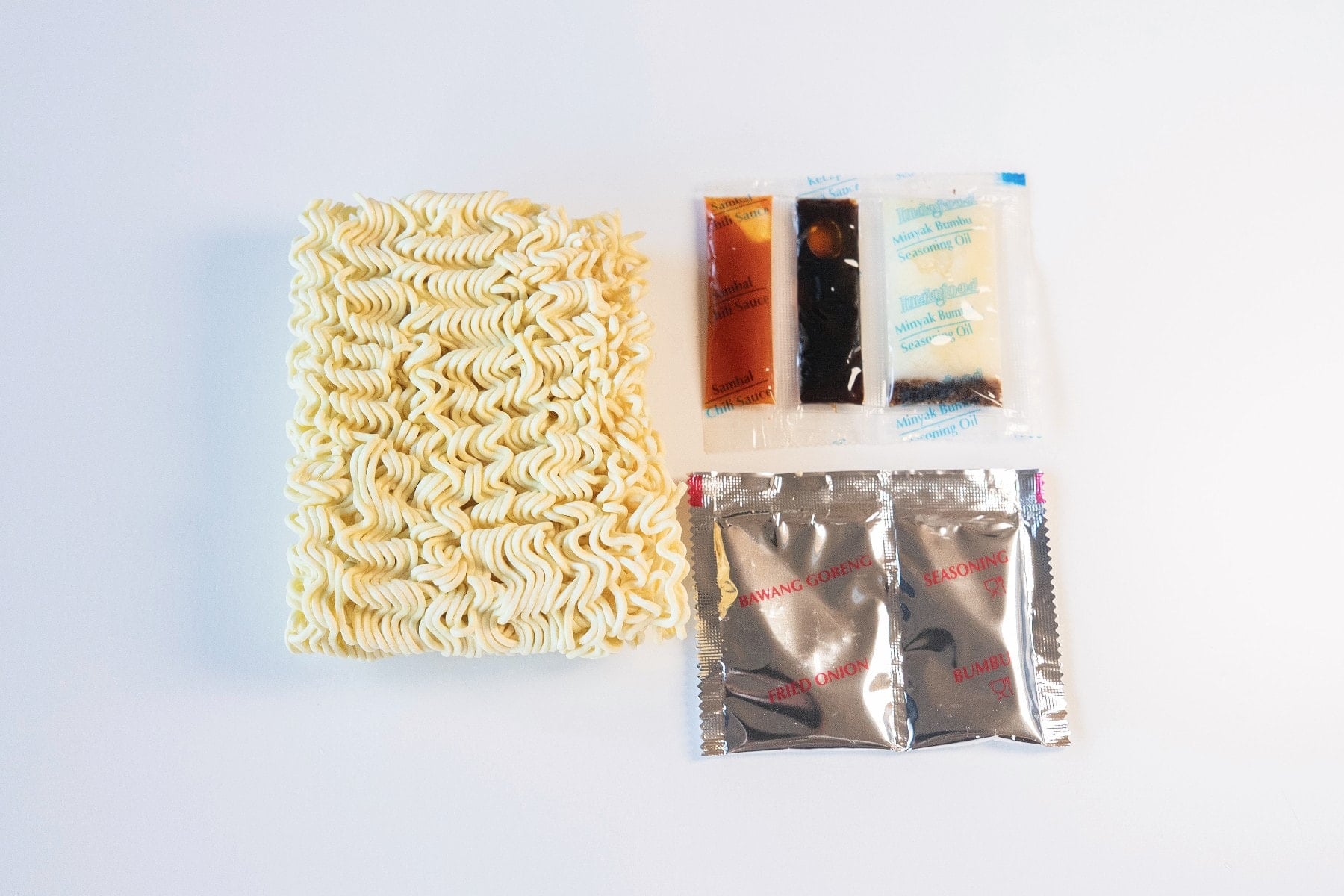 The contents of the Indomie Mi Goreng Instant Stir Fry Noodles pack laid out on a white surface, showing a rectangular block of thin wavy wheat noodles on the left with five sachets to the right. Three small horizontal sachets at top (orange, dark brown, clear) for the seasoning oil, sweet soy sauce, and chili sauce, and two silver foil sachets at the bottom labeled BAWANG GORENG (fried shallots) and SEASONING (bumbu).