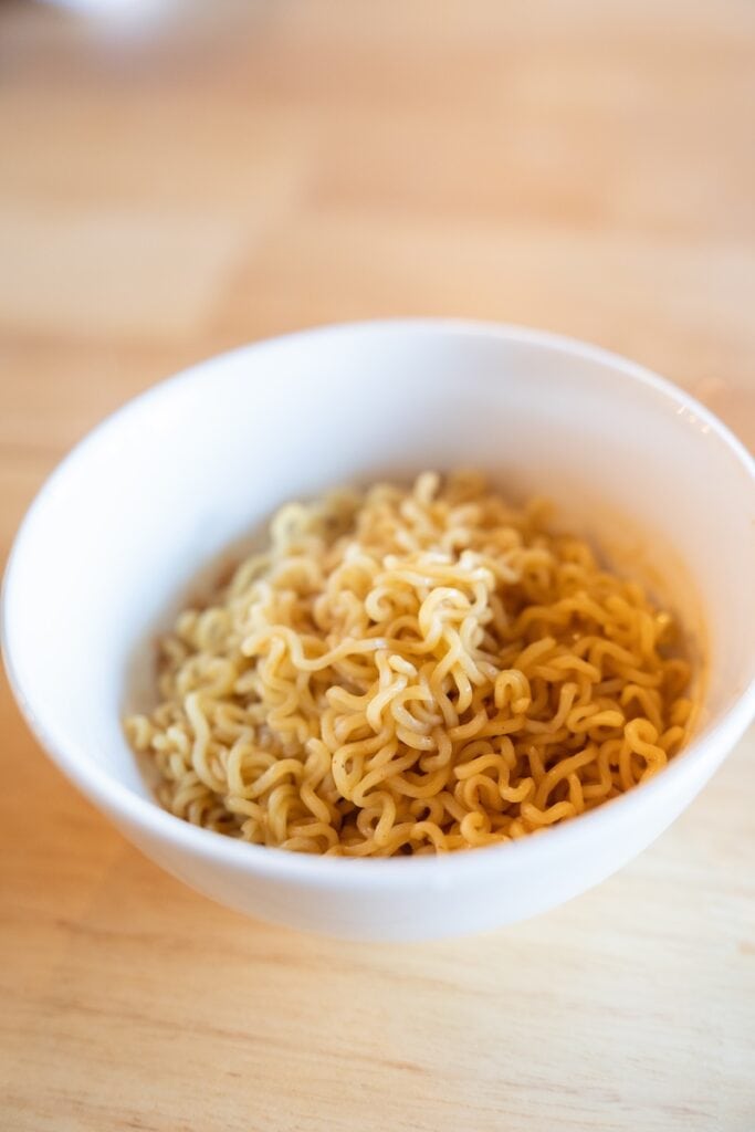 A white bowl of the finished Indomie Mi Goreng Instant Stir Fry Noodles on a wooden table, showing thin wavy wheat noodles coated in a glossy brown sweet soy sauce.