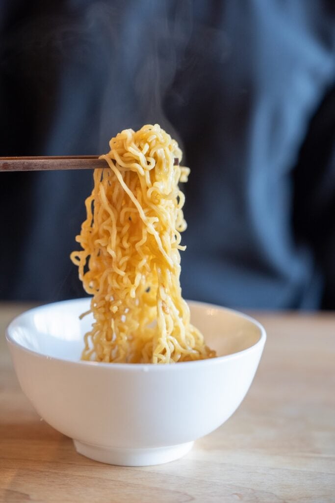 A close-up of the Indomie Mi Goreng Instant Stir Fry Noodles being lifted from a white bowl with wooden chopsticks, showing thin wavy wheat noodles coated in a glossy golden-brown sweet soy sauce with steam rising above them.