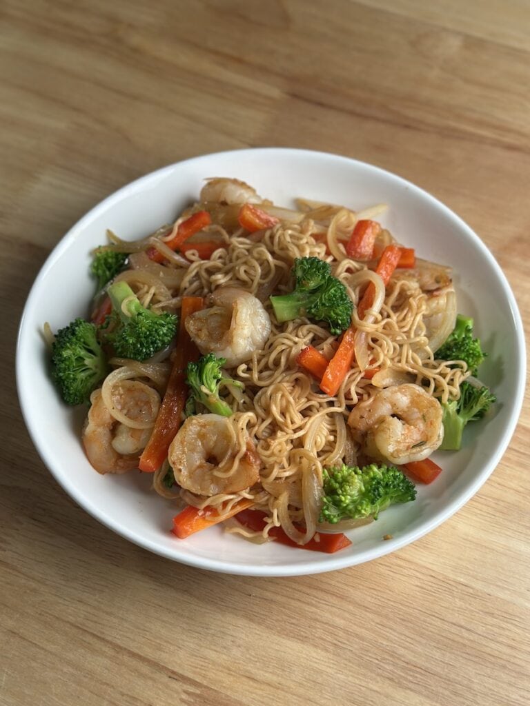 A white bowl filled with finished Instant Mi Xao Tom (Vietnamese stir-fried noodles with shrimp), featuring Hao Hao instant noodles, seared shrimp, bright green broccoli florets, red bell peppers, and onions, sitting on a wooden counter.