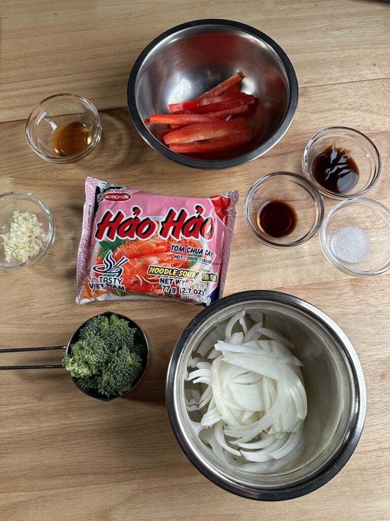 Ingredients for Instant Mi Xao Tom stir-fry including a pink package of Hao Hao Hot and Sour Shrimp instant noodles, sliced white onions, broccoli florets, sliced red bell peppers, minced garlic, and small bowls of sauce and seasoning.