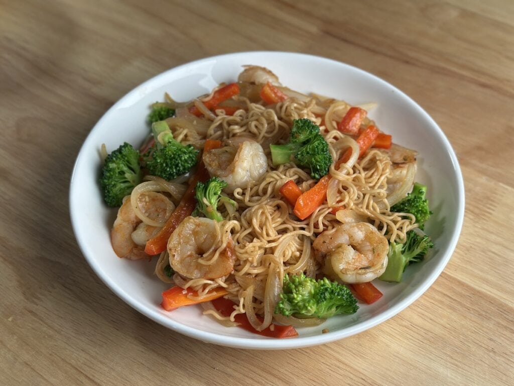 A white bowl filled with Instant Mi Xao Tom made with Hao Hao instant noodles, seared shrimp, broccoli, red bell peppers, and onions on a wooden table.