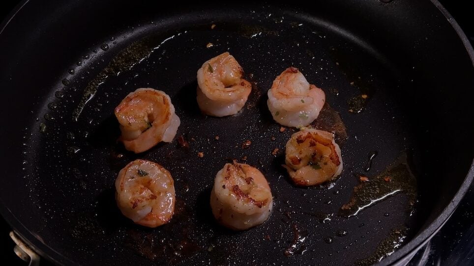 Six large, peeled shrimp searing in a hot, dark non-stick wok or skillet. The shrimp are turning pink and show a slight golden-brown char on the bottom.