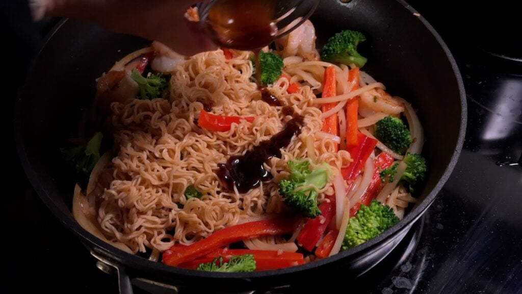 A hand pours a dark, savory stir-fry sauce from a small glass bowl over the Mi Xao Tom components in a hot wok. Visible ingredients include Hao Hao instant noodles, sliced red bell peppers, broccoli, and onions.