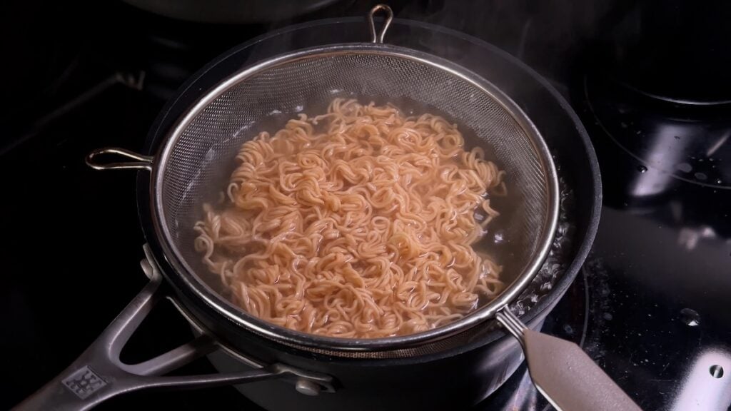 Dried instant ramen noodles soaking in hot, simmering water inside a metal mesh strainer nested within a black saucepan on a stovetop. Steam is rising from the water.