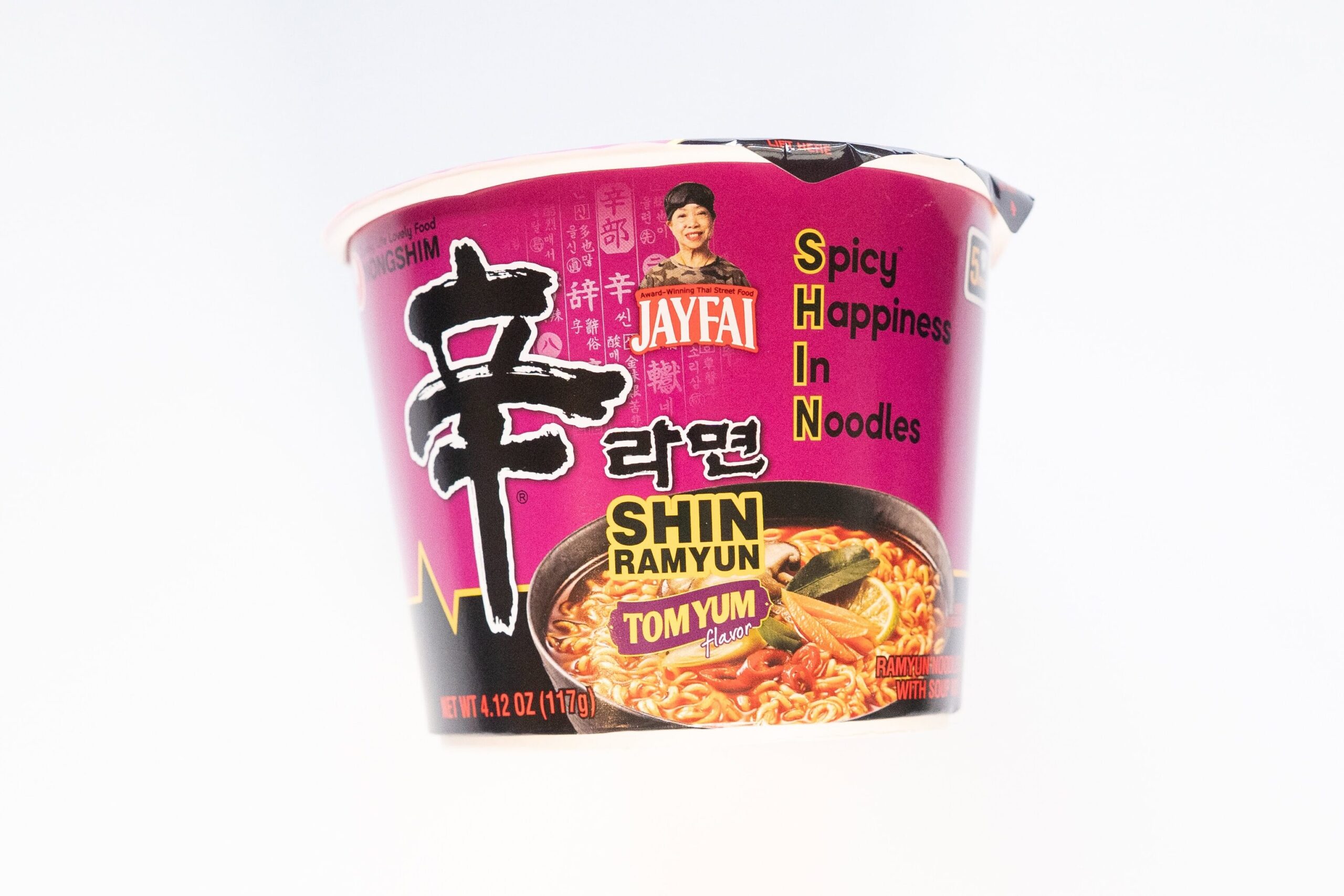 The deep magenta bowl of Nongshim Shin Ramyun Tom Yum Flavor Jay Fai edition on a white surface, showing the white NONGSHIM logo, the large hand-brushed black 辛 character, SHIN RAMYUN in black and yellow text, a purple TOM YUM flavor banner, the JAYFAI yellow logo with Jay Fai's photo, SPICY HAPPINESS IN NOODLES vertical text down the right side, and a photo of the finished Tom Yum ramyun in a black bowl with chili and kaffir lime leaf.