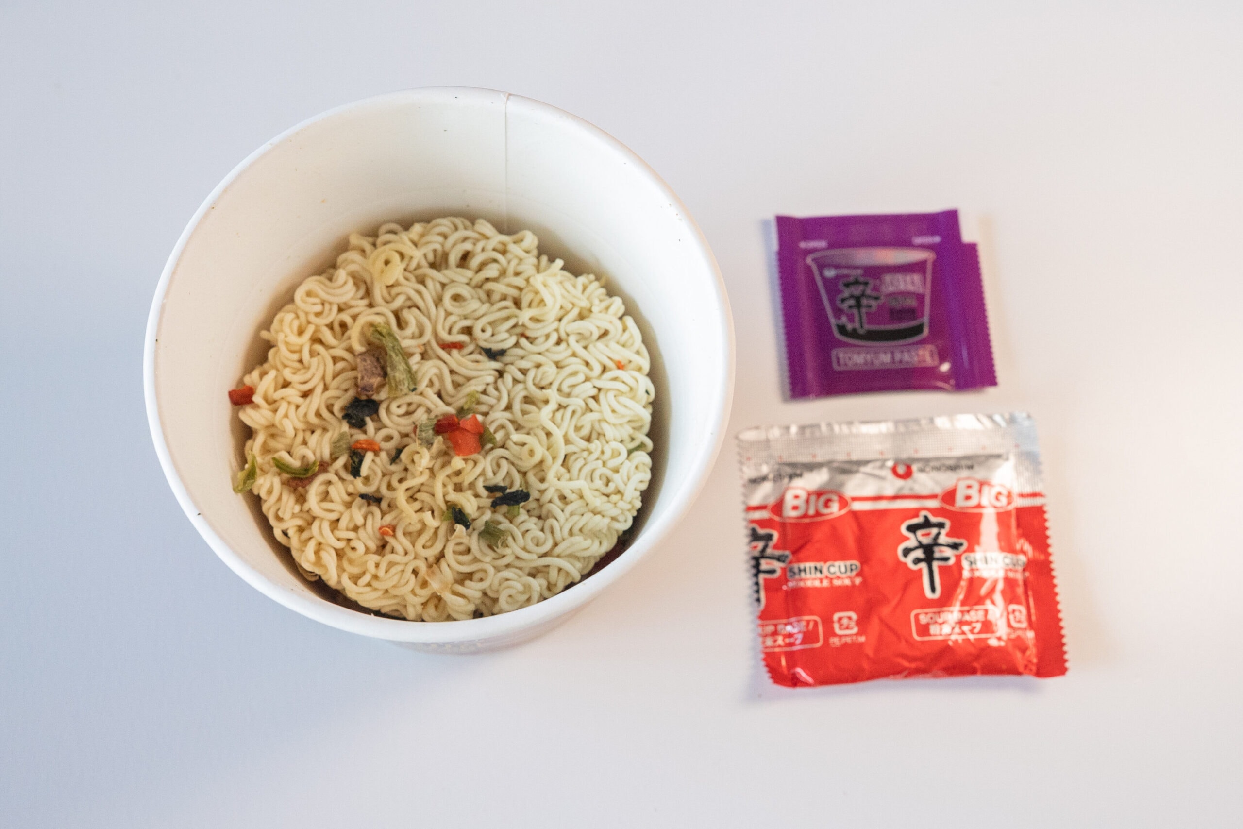 The contents of the Nongshim Shin Ramyun Tom Yum Flavor Jay Fai cup laid out on a white surface, showing the empty paper bowl with a round disc of fried wavy wheat noodles dotted with dehydrated vegetable flecks, a purple TOM YUM PASTE sachet, and a red Shin Cup SOUP BASE packet.