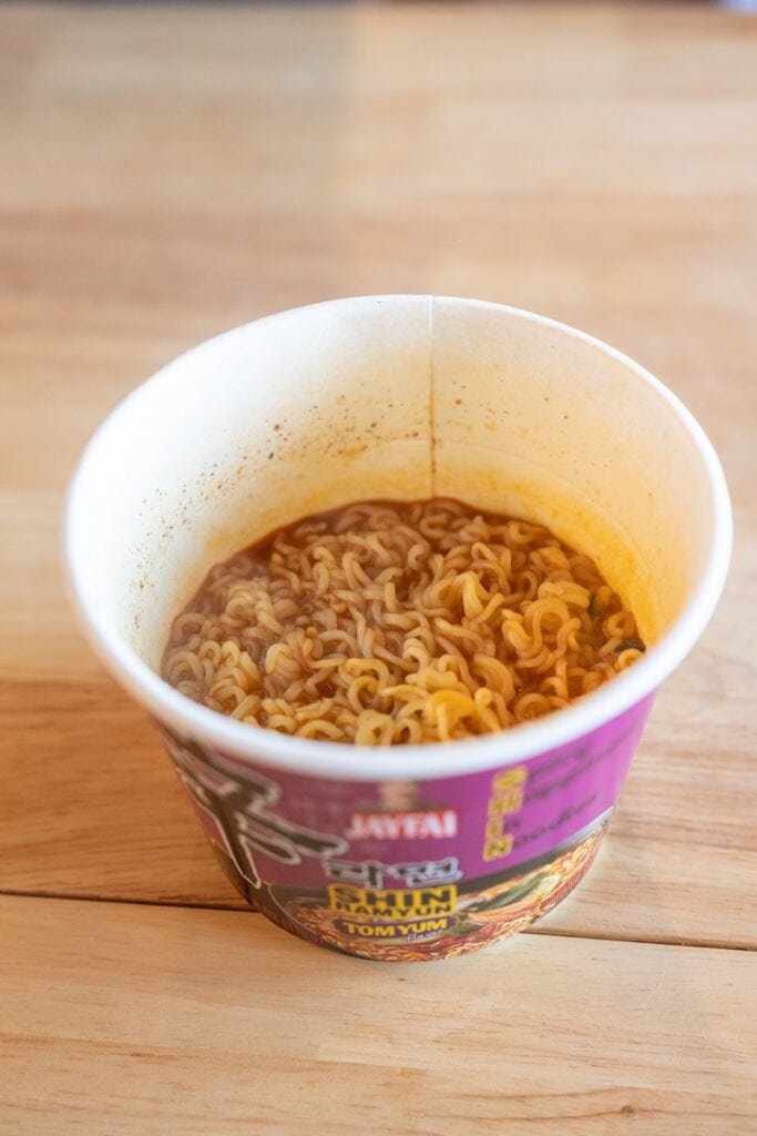 A three-quarter view of the finished Nongshim Shin Ramyun Tom Yum Flavor Jay Fai in its paper cup, showing wavy wheat noodles in a deep red-brown Tom Yum broth with visible green onion flecks floating on top.