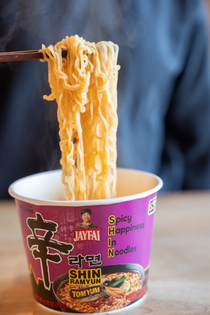 A close-up of the Nongshim Shin Ramyun Tom Yum Flavor Jay Fai being lifted from the paper cup with wooden chopsticks, showing wavy wheat noodles glossy with red-orange Tom Yum broth and steam rising above them.