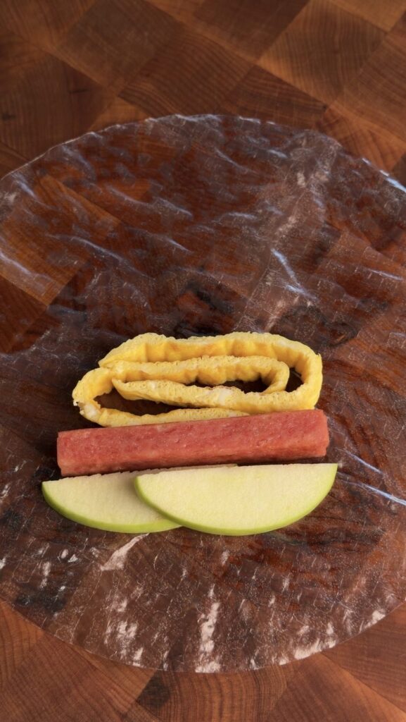 A rice paper wrapper laid out with egg ribbons, a Spam strip, and green apple slices placed for assembly