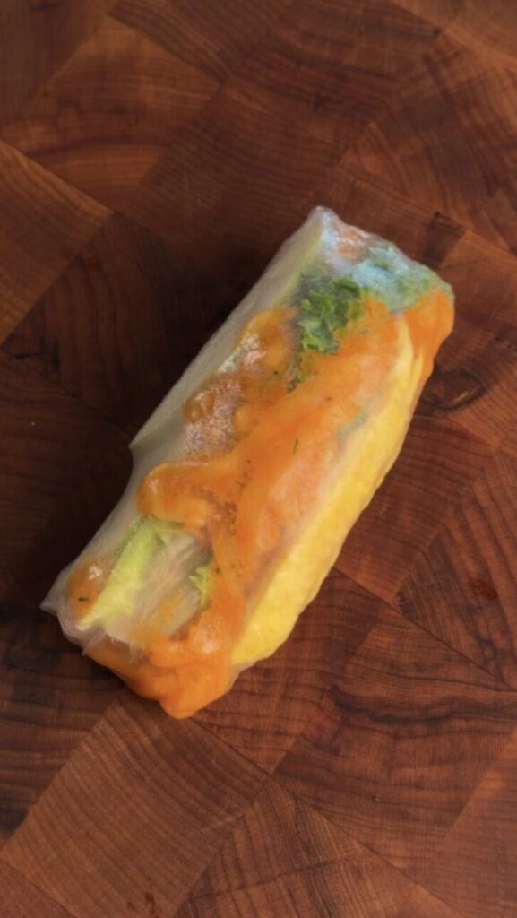 One whole Buldak Carbonara spring roll on a wood cutting board before slicing, with lettuce and creamy noodles visible through the rice paper