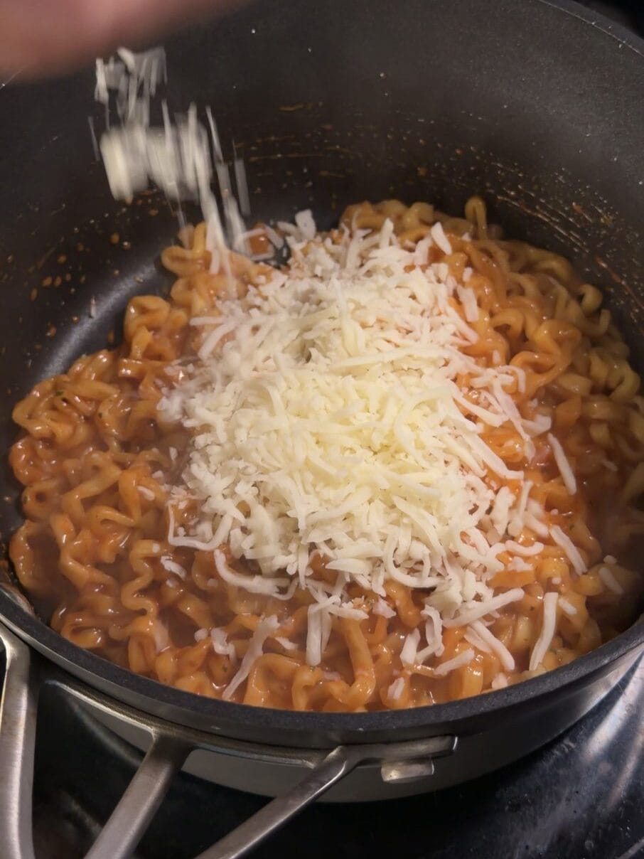 A handful of shredded fresh mozzarella dropped onto Buldak Carbonara noodles in a pan to build the creamy sauce