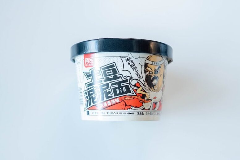 The white and red cup of A-Kuan Instant Mashed Potato Noodles (Tomato Flavor) on a white surface, showing a small red 阿宽 (A-Kuan) logo at the top left, the large black Chinese characters 土豆泥泥面 (Mashed Potato Mud Noodles), a 浓香番茄味 (Strong Tomato Flavor) tag, an illustration of a man wearing a chef-style helmet eating noodles, the romanized name TU DOU NI NI MIAN, and a black plastic lid on top.