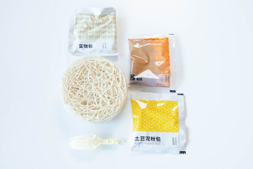 The contents of the A-Kuan Instant Mashed Potato Noodles cup laid out on a white surface, showing a round nest of thin straight wheat noodles in the center, a small silver foil ๅฎ็ฉๅ
(Solid Pack) sachet at the top with dehydrated bits visible, a red ้
ฑๅ
(Sauce) pouch on the right with orange-red tomato sauce, a yellow ๅ่ฑๆณฅ็ฒๅ
(Potato Powder Pack) at the bottom right, and a small white plastic spork at the bottom.
