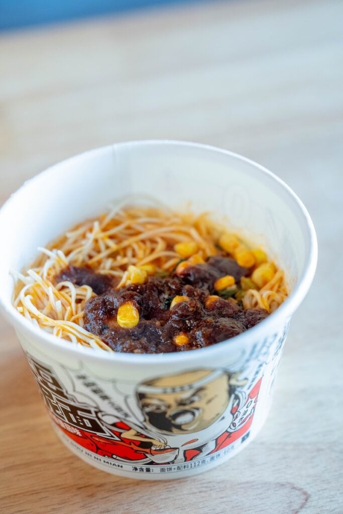 A top-down view of the finished A-Kuan Instant Mashed Potato Noodles in the paper cup, showing thin straight wheat noodles fully coated in a thick orange-red tomato-and-mashed-potato sauce with visible green herb flecks and small corn kernels.