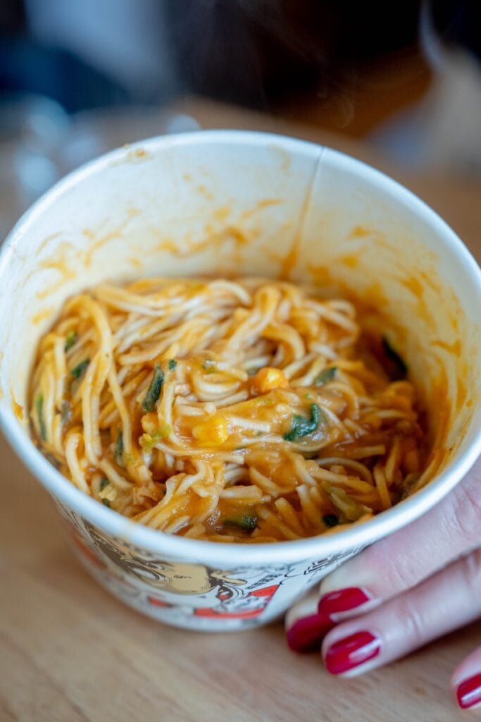 A top-down view of the finished and mixed A-Kuan Instant Mashed Potato Noodles in the paper cup, showing thin straight wheat noodles fully coated in a thick orange-red tomato-and-mashed-potato sauce with visible green herb flecks and small corn kernels.
