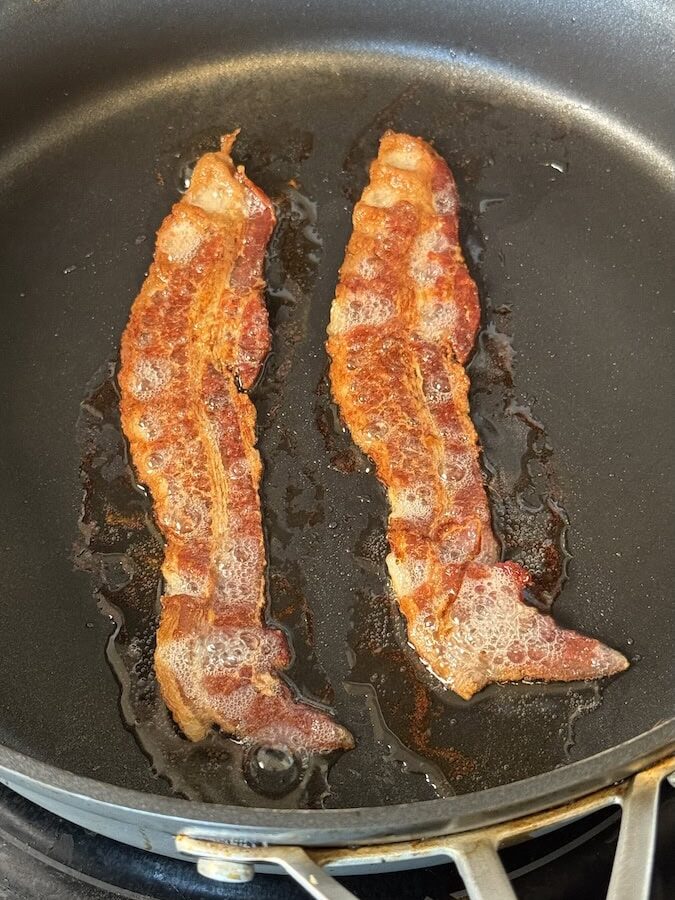 Two large, crispy strips of fried bacon cooking in a black non-stick skillet.