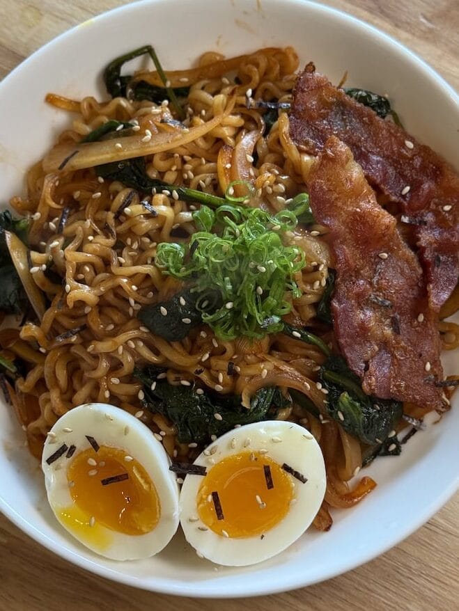 Bowl of noodles topped with pan-fried bacon, spinach, soft-boiled egg, green onions, and sesame seeds
