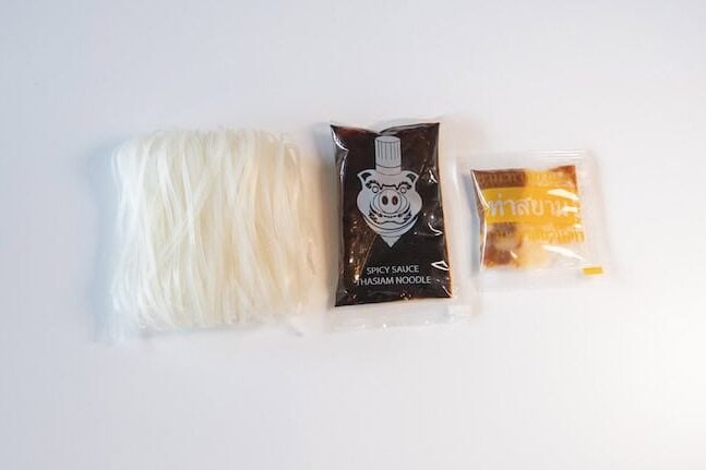 The contents of the Thasiam Instant Dried Rice Stick Noodles with Spicy Sauce pack laid out on a white surface, showing a bundle of thin white dried rice stick noodles on the left, a dark Spicy Sauce pouch with a small pig chef illustration in the middle, and a small clear yellow seasoning sachet on the right.