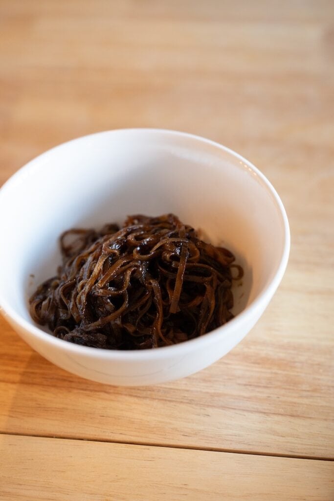 A white bowl of the finished Thasiam Instant Dried Rice Stick Noodles with Spicy Sauce on a wooden table, showing thin rice stick noodles coated in a dark glossy spicy-sour sauce.