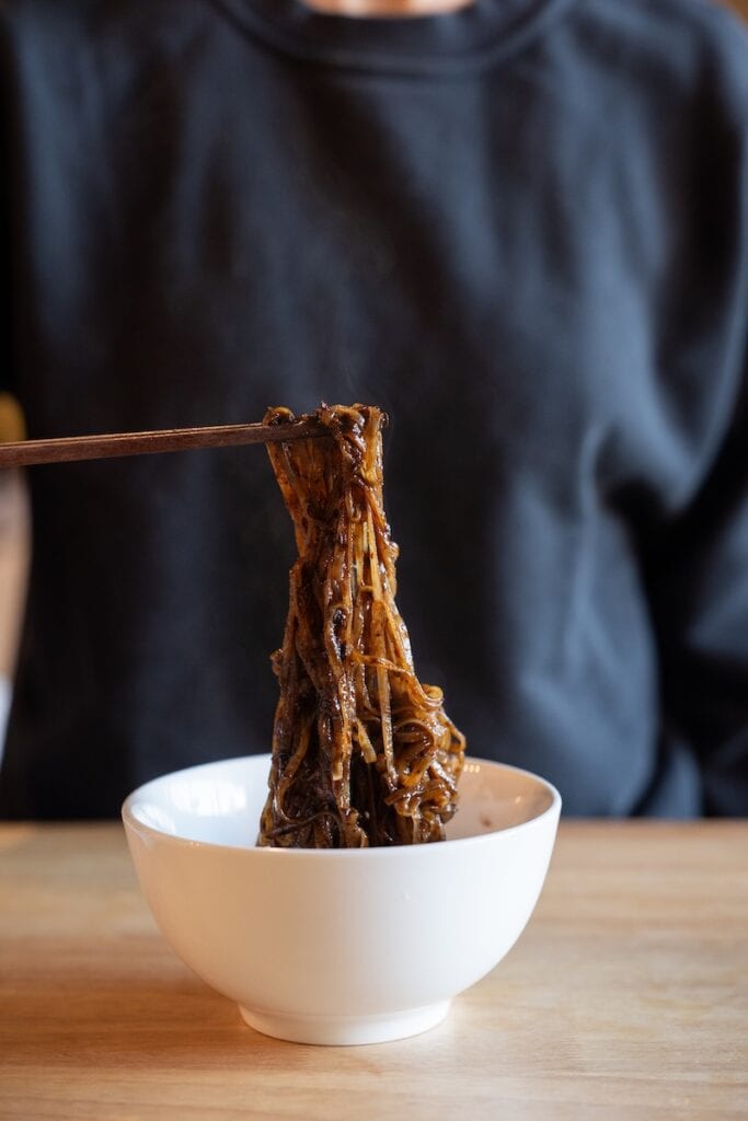 A close-up of the Thasiam Instant Dried Rice Stick Noodles with Spicy Sauce being lifted from a white bowl with wooden chopsticks, showing thin rice stick noodles glossy with a dark brown-black spicy sauce and steam rising above them.