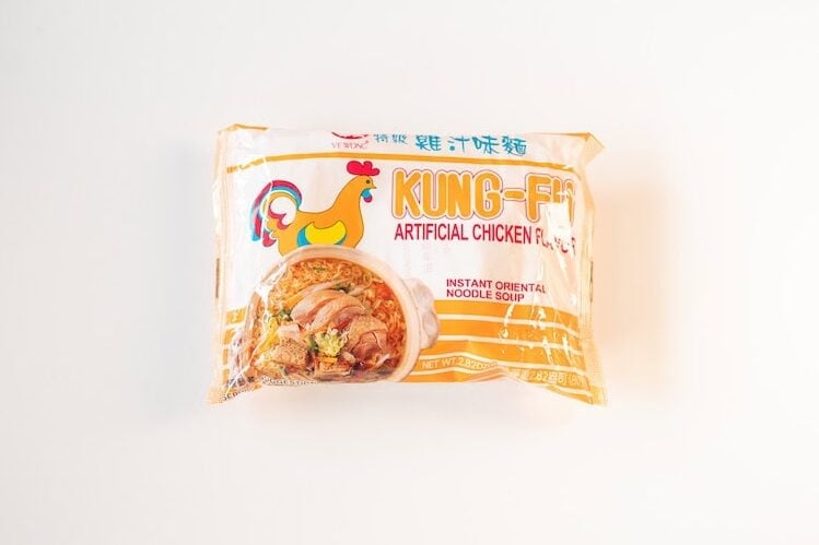 The cream and yellow package of Ve Wong Kung-Fu Chicken Flavor Instant Noodle on a white surface, showing a small yellow cartoon rooster on the left, KUNG-FU in large red lettering, ARTIFICIAL CHICKEN FLAVOR in red below, INSTANT ORIENTAL NOODLE SOUP on the right side, and a photo of the finished noodles in a small ceramic bowl.