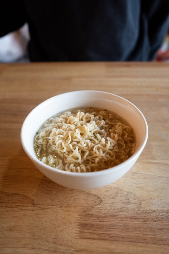 A white bowl of the finished Ve Wong Kung-Fu Chicken Flavor Instant Noodle on a wooden table, showing thin wavy pale wheat noodles in a clear light chicken broth with faint green herb specks.
